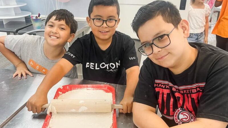 Three young chefs gather around a rolling pin and floured dough at Flour Power Studios.